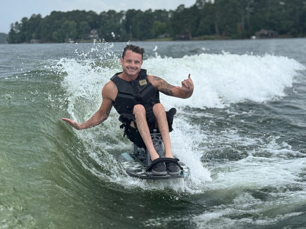 A smiling man wearing a black life vest rides an adaptive seated wakesurf board on a lake wave. He balances with his right hand extended and flashes a shaka sign with his left hand amidst water spray, with a treeline visible in the far background.