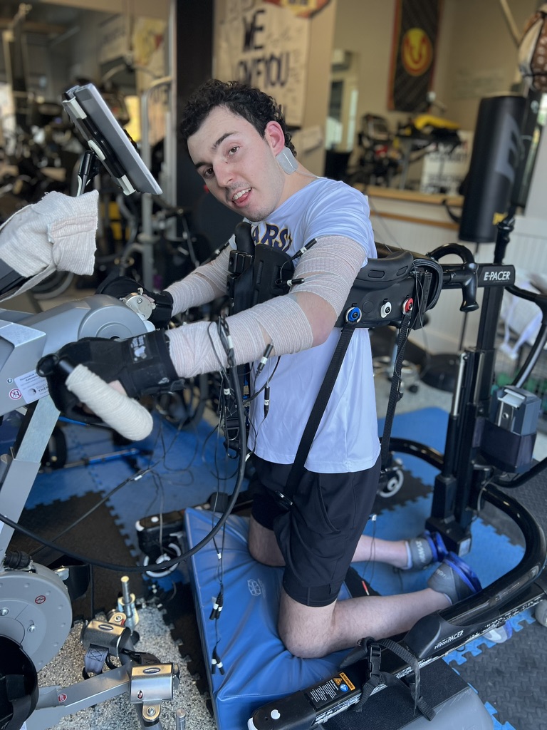 Devon Corbett kneels on a blue foam mat while supported by a black E-Pacer mobility frame, gripping the hand controls of a grey functional electrical stimulation (FES) therapy machine. His arms are wrapped in medical bandages with electrodes attached, and he looks toward the camera with a playful expression inside a gym-style rehabilitation facility.