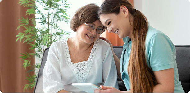 A panoramic medium shot of an older woman with glasses and a white lace-trimmed top sitting on a sofa next to a young female nurse or caregiver wearing a stethoscope. Both are smiling warmly and leaning close together as they look down at a digital tablet held by the caregiver.