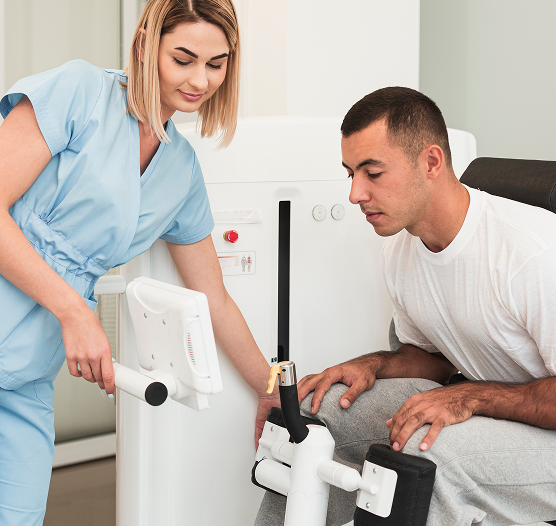 A medium shot of a female physical therapist in light blue scrubs leaning forward to adjust a digital screen connected to an advanced leg rehabilitation machine. A male patient in a white t-shirt and grey sweatpants sits on the machine, looking intently at his knee as it is positioned within the padded supports of the device.
