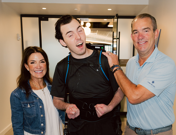 Devon Corbett stands in a hallway supported by a robotic exoskeleton, smiling alongside his parents who stand on either side of him in support.