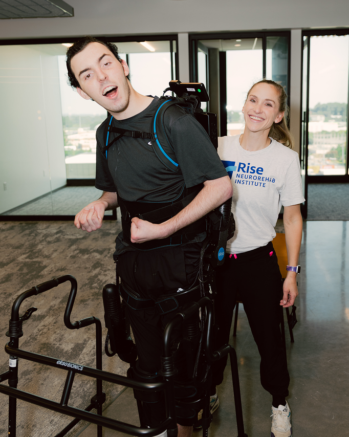 Devon Corbett stands using a black wheeled walker while wearing a robotic exoskeleton device. Behind him, his therapist Caitlyn, wearing a 'Rise Neurorehab Institute' t-shirt, stands smiling broadly in support inside a bright therapy room with glass windows.