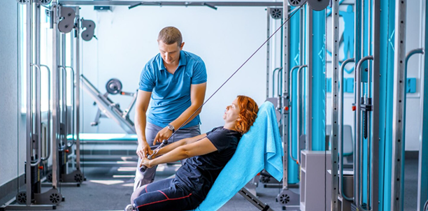 A panoramic medium shot of a woman with red hair sitting on an inclined workout bench with a blue towel behind her head, performing a cable pulldown exercise. A male physical therapist or personal trainer in a blue polo shirt stands beside her, guiding her form in a brightly lit gym or rehabilitation facility.