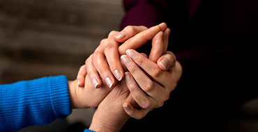 A close-up photograph capturing a supportive gesture where one person gently clasps another person's hand with both of theirs. A person wearing a blue ribbed sleeve holds an older individual's hand, while a second set of hands rests comfortingly on top against a dark, blurred background.