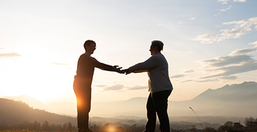 A panoramic wide silhouette shot of two individuals facing each other and holding hands in a field at sunrise or sunset. The golden sun low on the horizon casts a bright glow over a sprawling valley and distant mountains in the background.
