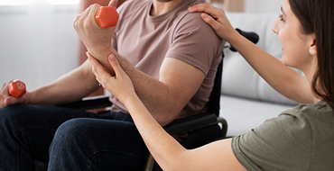 A close-up shot of a man sitting in a wheelchair, lifting a small orange dumbbell during a physical therapy or rehabilitation session. A female therapist or caregiver stands beside him, smiling as she places one hand on his shoulder and gently guides his forearm to support his movement.