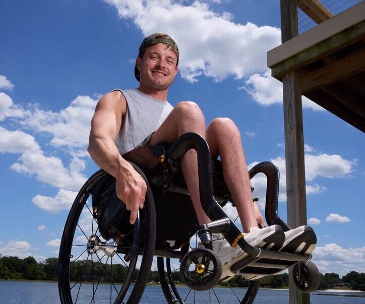 A low-angle photograph of a smiling young man in a backwards baseball cap and grey sleeveless shirt performing a wheelie in his manual wheelchair on an outdoor wooden dock. The background features a calm body of water, trees, and a bright blue sky filled with white clouds.