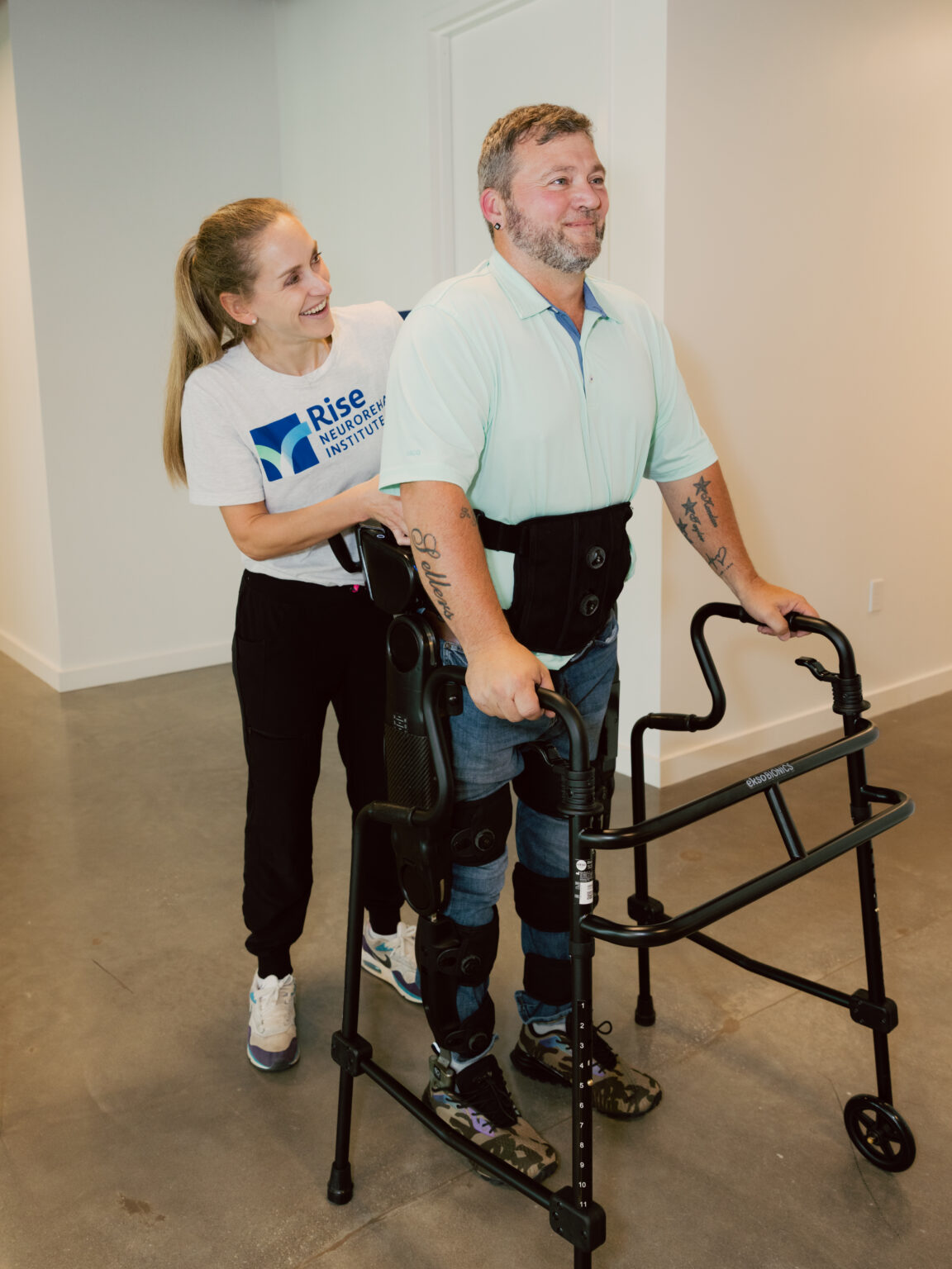 A man named Bull Sellers stands in an indoor hallway, supported by a black robotic exoskeleton and holding onto a walker while looking forward. Behind him, therapist Caitlyn, wearing a 'Rise Neurorehab Institute' t-shirt, smiles warmly while supporting the exoskeleton's harness from behind.