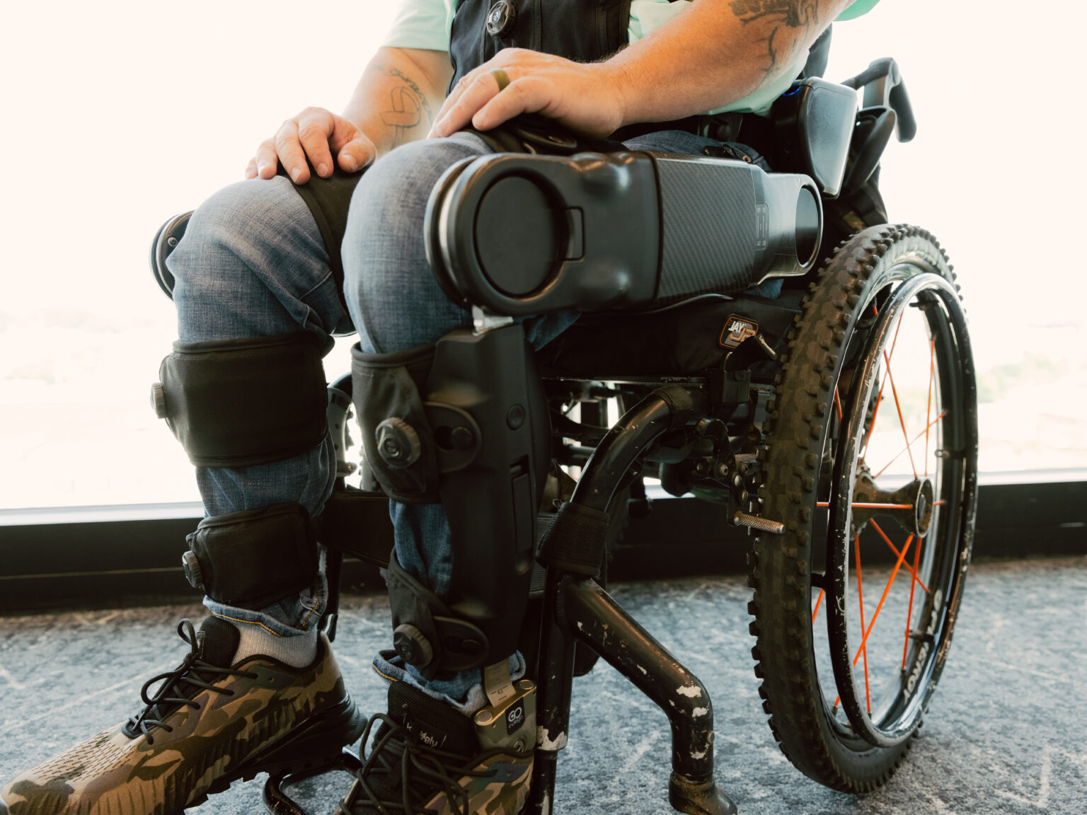A close-up of a man sitting in a wheelchair, wearing a black robotic exoskeleton device strapped tightly over his legs and jeans. The wheelchair features knobby tires with orange spokes, and a bright window is visible in the background.