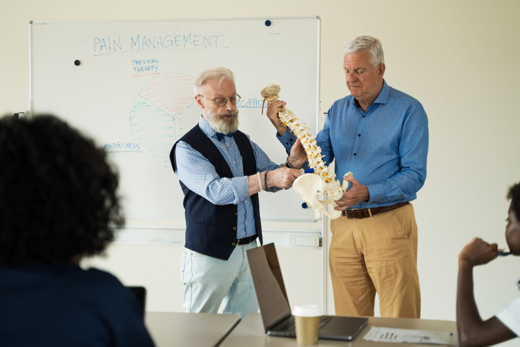 Senior medical experts presenting spine model during pain management seminar. Whiteboard shows holistic approach including physical therapy, medication, and mindfulness. Diverse audience attends.