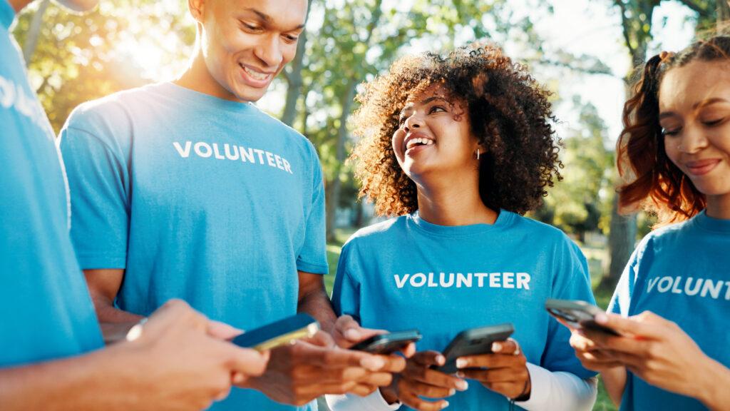 Volunteers, charity and team with phone, outdoor and communication for fundraising event or people. Happy, social responsibility and group with tech for donations, browsing and community service.