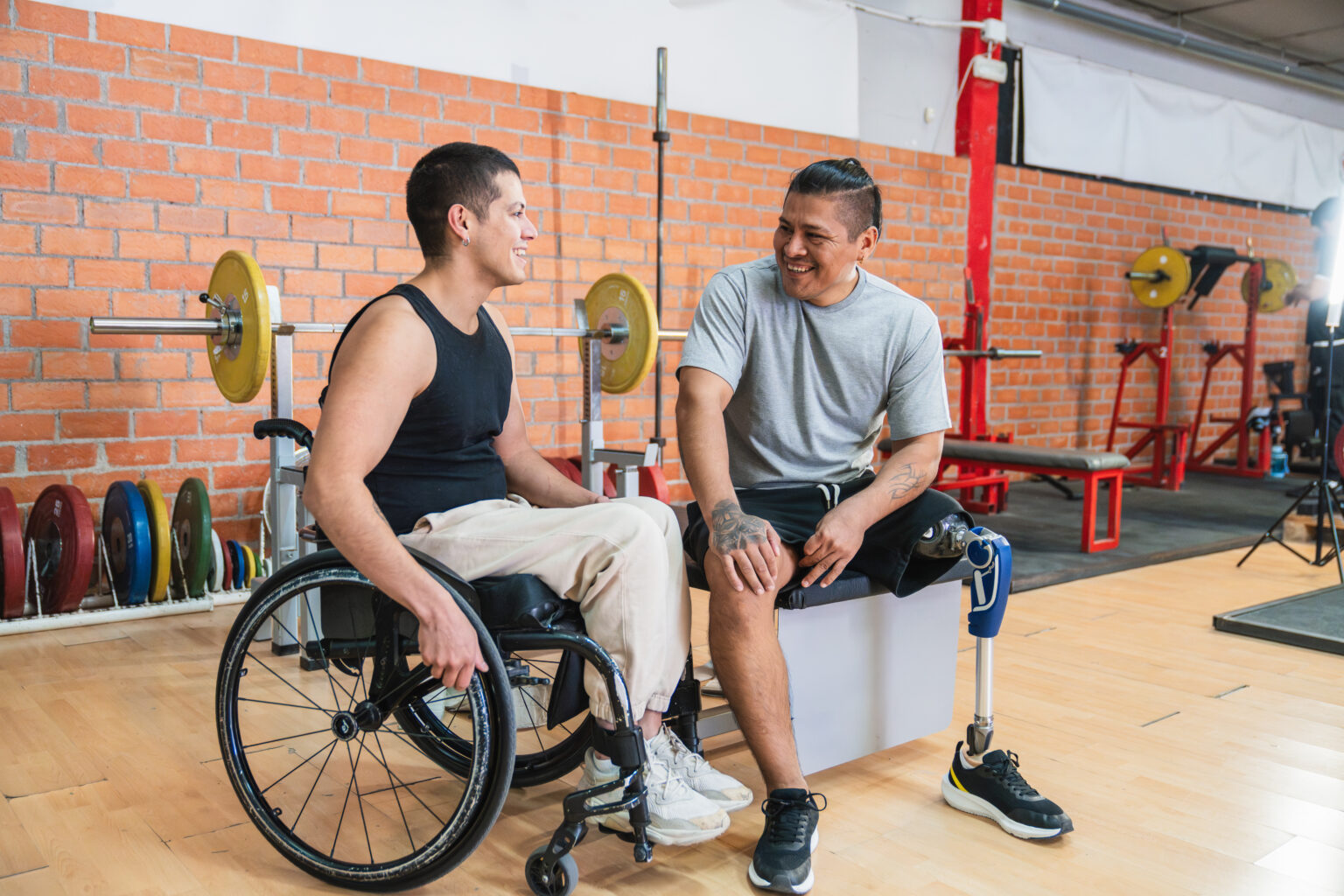 Two young disabled friends, one in a wheelchair and one with a prosthetic leg, smiling and talking during a gym session
