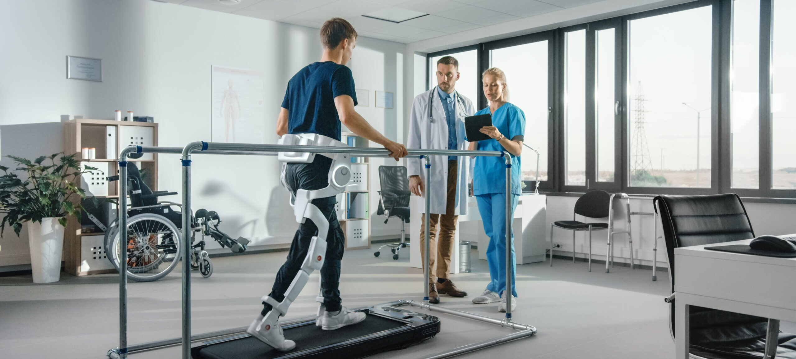 Modern Hospital Physical Therapy: Patient with Injury Walks on Treadmill Wearing Advanced Robotic Exoskeleton. Physiotherapy Rehabilitation Scientists, Engineers, Doctors use Tablet Computer to Help